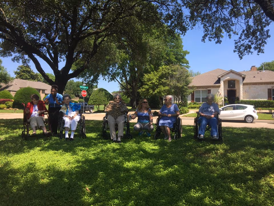 A group of six elderly women, five in wheelchairs and one sitting on the grass, are gathered outside on a sunny day under the shade of a large tree. They are on a grassy area in front of residential houses with a clear blue sky above.