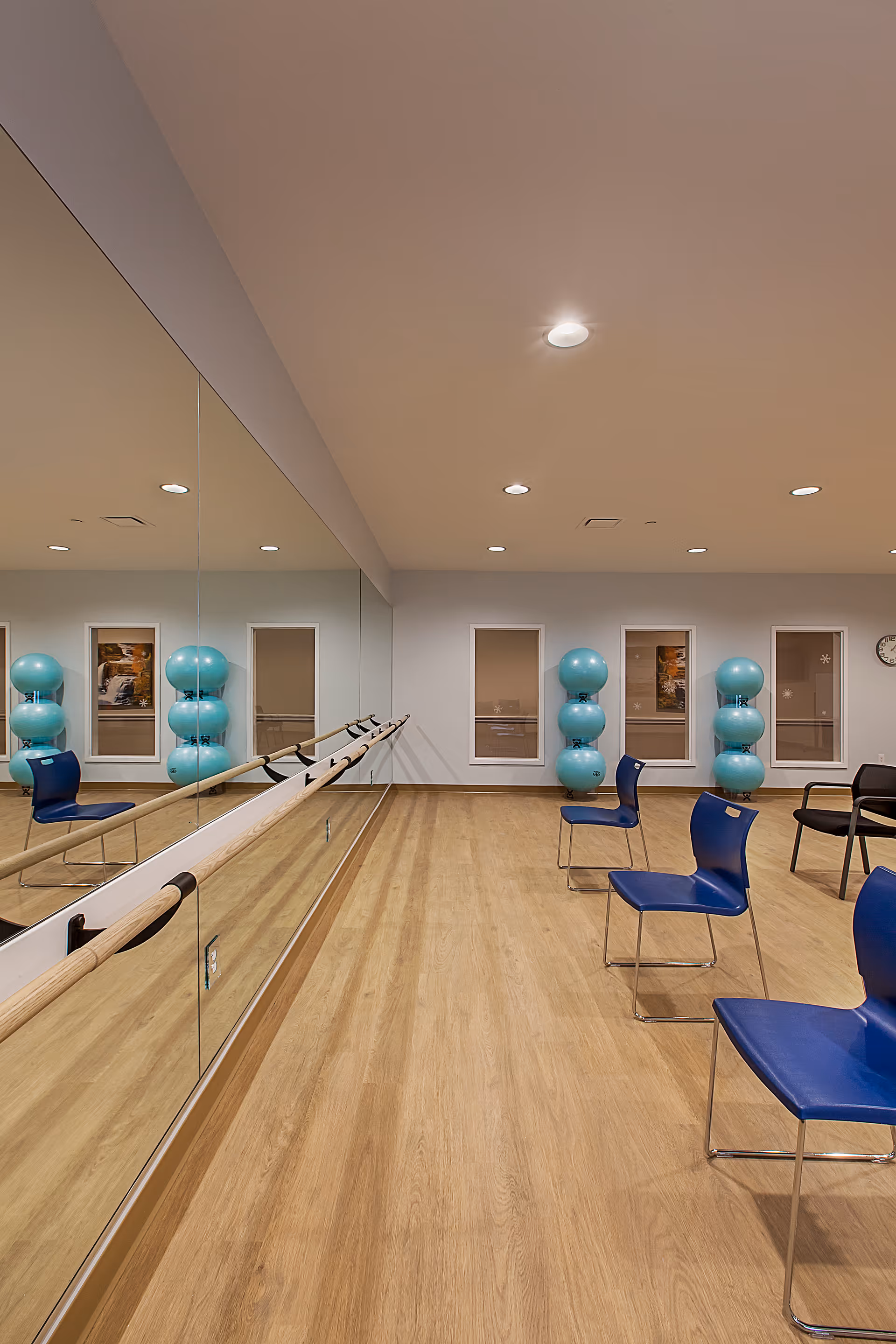 A spacious exercise room with wooden flooring, a wall-length mirror with ballet barres, blue chairs arranged in rows, and stacks of large blue exercise balls against the far wall under three windows.