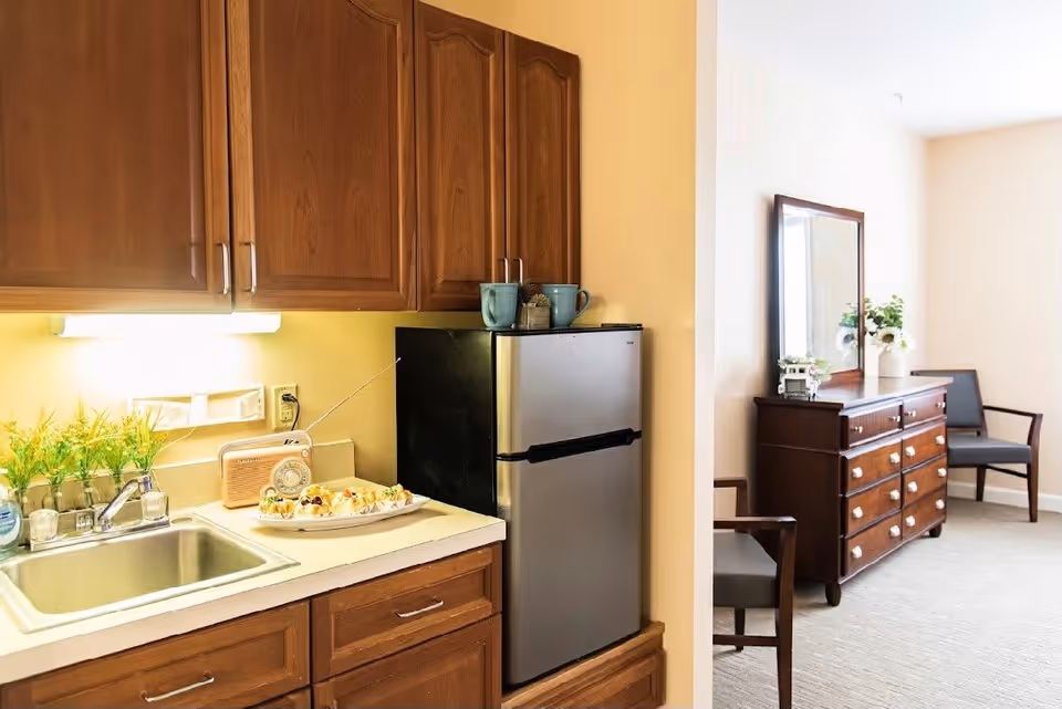 A small kitchen area with wooden cabinets, a stainless steel mini refrigerator, a sink, and a countertop with a plate of pastries and a vintage radio. Adjacent to the kitchen is a room with a wooden dresser, a large mirror, two chairs, and a vase with flowers.