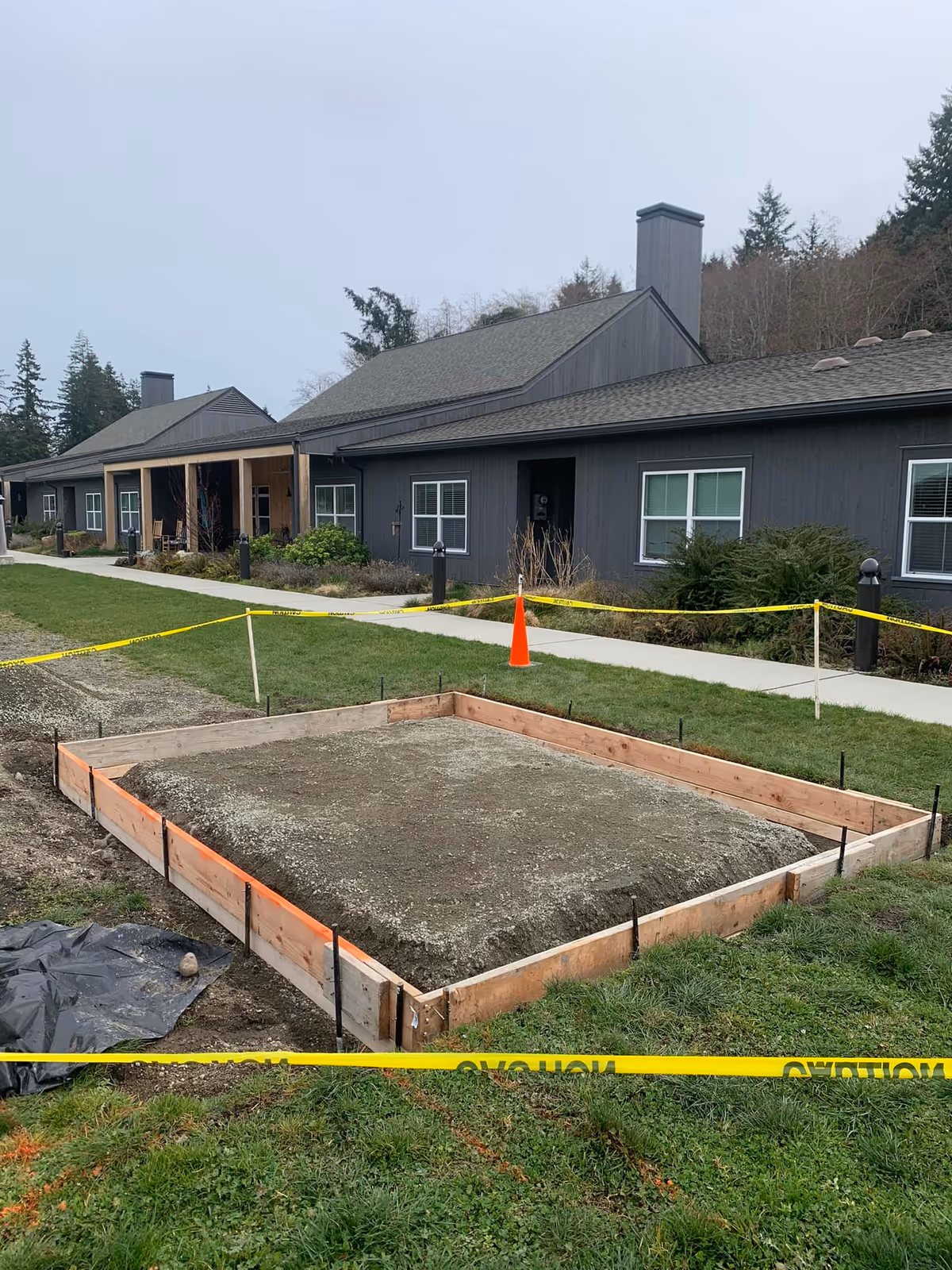 Construction site with a wooden frame outlining a square area filled with gravel, surrounded by yellow caution tape and an orange traffic cone. In the background, there is a dark gray building with multiple windows and a covered porch, set against a backdrop of trees and a cloudy sky.