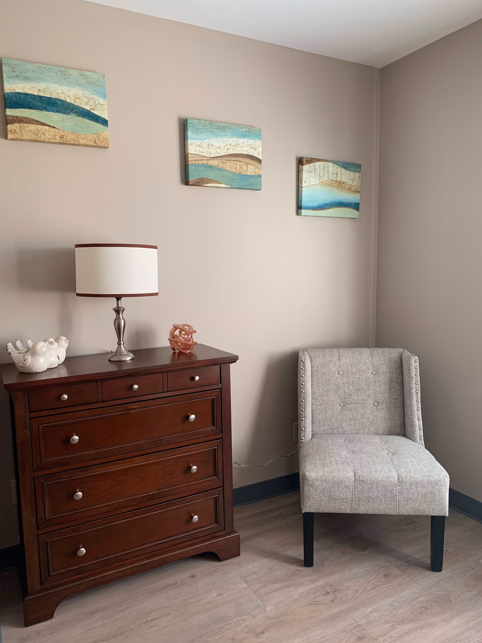 A corner of a room featuring a dark wooden chest of drawers with a white table lamp, a ceramic bird decoration, and a small decorative object on top. Next to the chest is a light gray upholstered chair with button tufting and nailhead trim. Three abstract landscape paintings with blue, beige, and brown tones hang on the beige wall above the furniture. The floor is light wood.