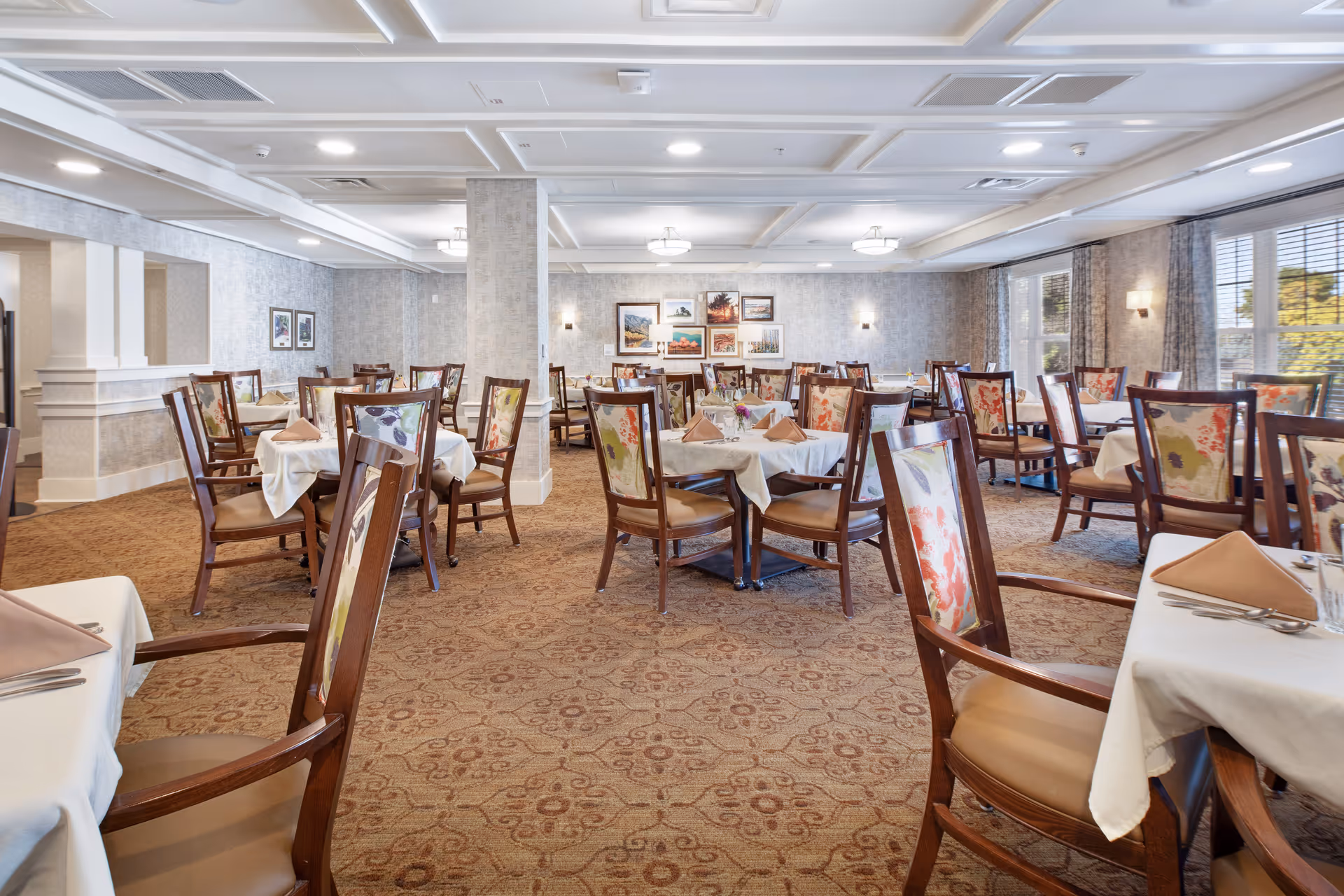 A spacious dining room with multiple tables covered in white tablecloths and set with brown napkins and silverware. The chairs have wooden frames with floral patterned upholstery. The room has beige patterned carpet, light-colored walls with framed pictures, and large windows with curtains allowing natural light to enter.