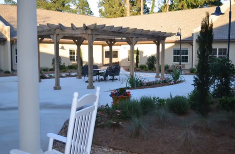 Outdoor courtyard area at Cedar Crest Memory Care featuring a wooden pergola with a table and chairs underneath, surrounded by landscaped garden beds with flowers and shrubs. A white rocking chair is visible in the foreground, and the building with windows and doors encloses the courtyard.