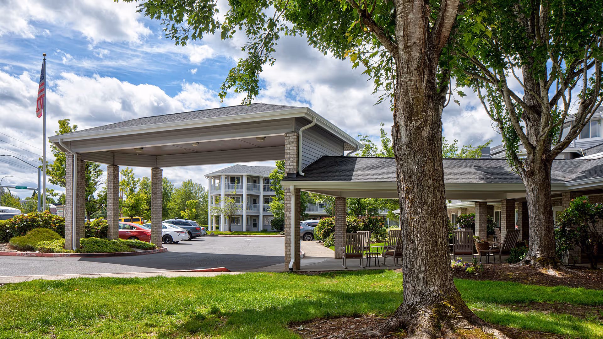 Entrance area of Holiday Rock Creek facility with a covered drop-off zone supported by brick pillars, surrounded by green grass, trees, and outdoor seating. Several cars are parked in the background under a partly cloudy sky.