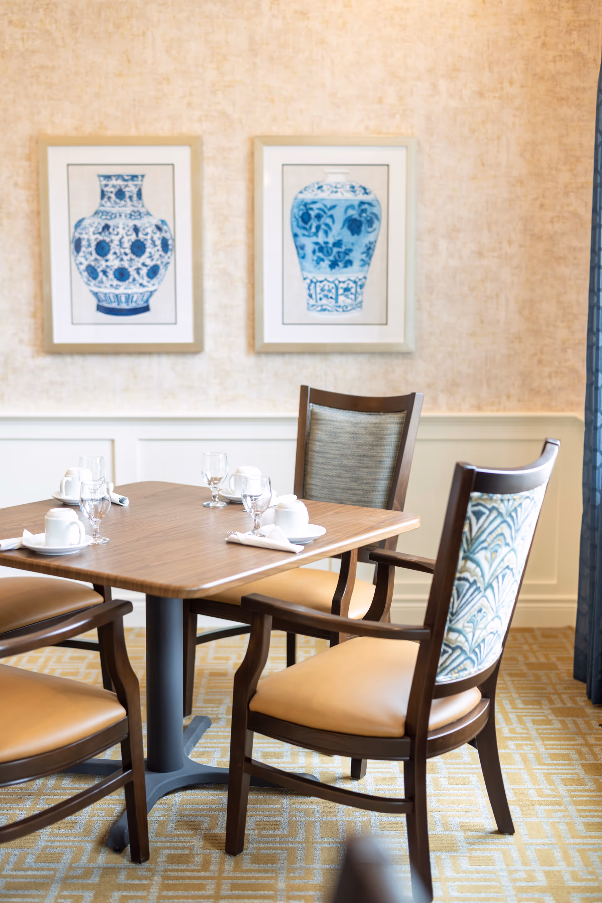 A dining table set for four with wooden chairs featuring cushioned seats and patterned backs. The table has white cups, saucers, napkins, and glasses. Two framed blue and white vase artworks hang on the beige textured wall behind the table.
