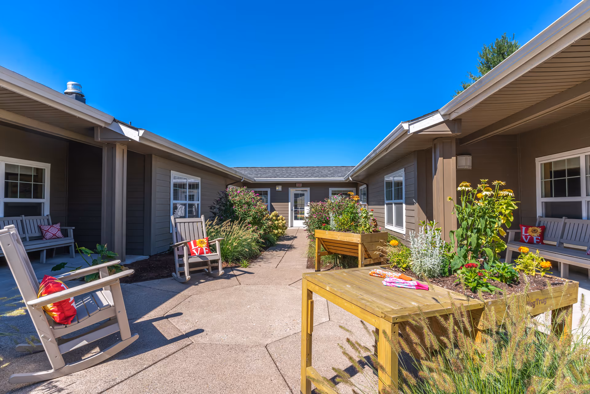 Outdoor courtyard area at Evergreen Memory Care with rocking chairs featuring red cushions, raised garden beds with various plants and flowers, and a clear blue sky overhead. The courtyard is surrounded by single-story buildings with windows and benches.