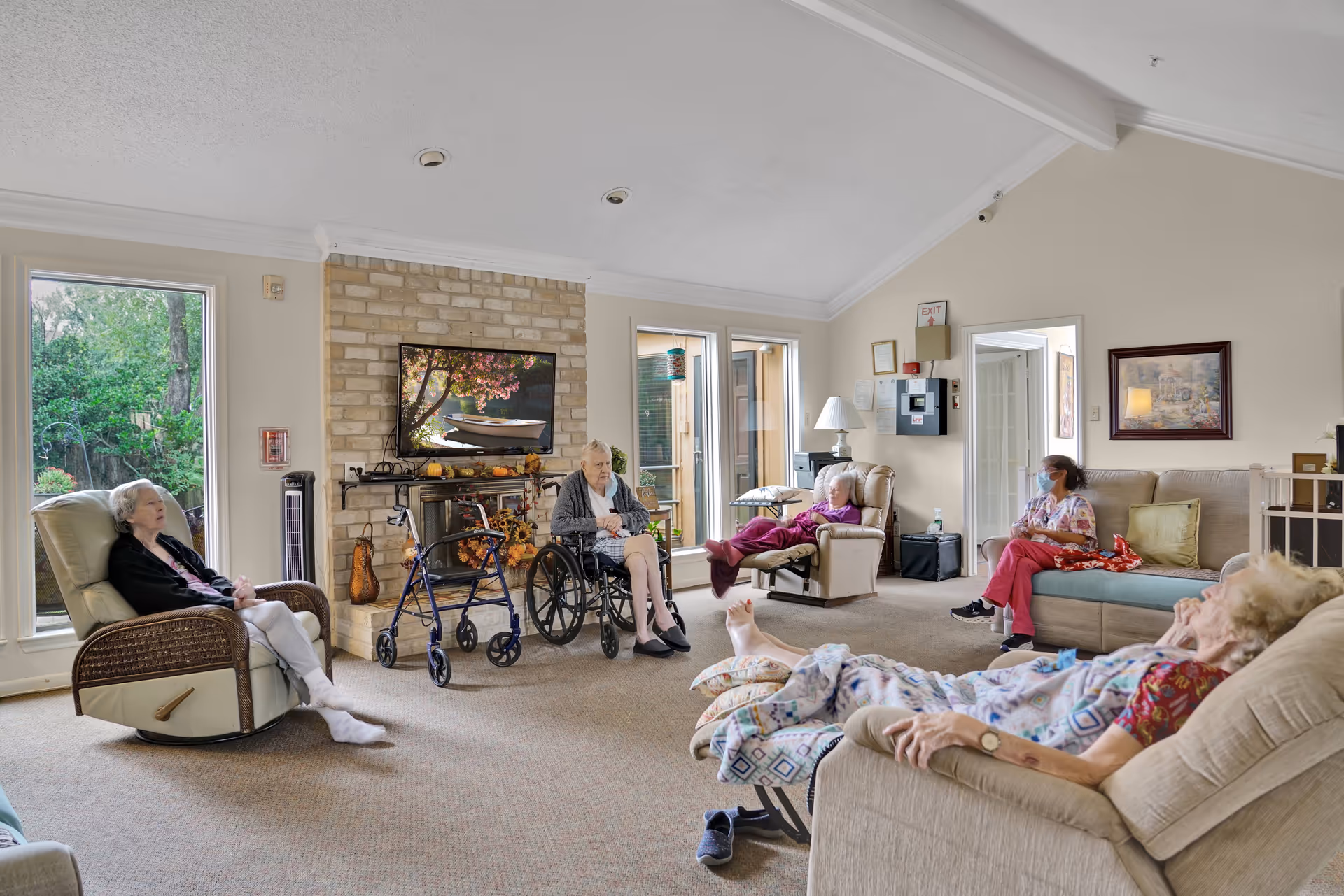A bright communal living room in a memory care facility with elderly residents seated in recliners and a wheelchair facing a wall-mounted TV over a brick fireplace.