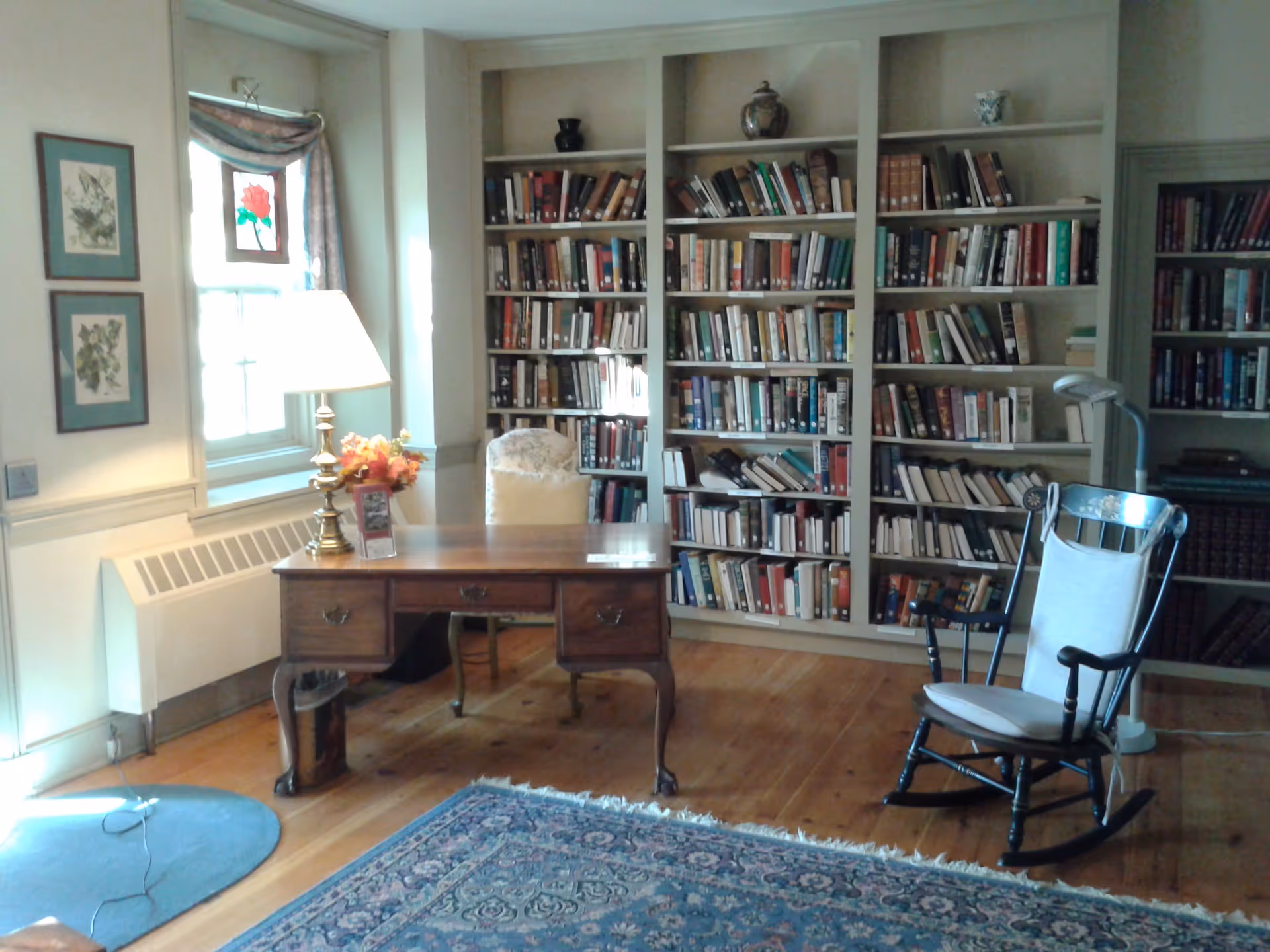 A cozy room with wooden flooring featuring a large bookshelf filled with books against the wall. In front of the bookshelf is a wooden desk with a lamp and a flower arrangement on it. To the right of the desk is a wooden rocking chair with a white cushion. There is a window with a floral curtain and framed botanical prints on the wall to the left. A blue patterned rug covers part of the floor.