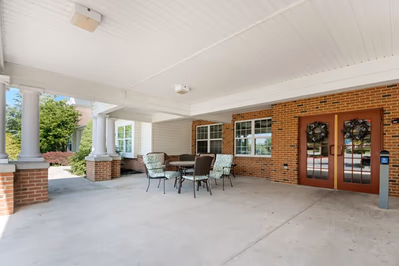 Covered outdoor seating area with a round table and four cushioned chairs under a white ceiling supported by brick and white columns. There are two red double doors with wreaths on them and windows on the brick wall behind the seating area. Greenery and a pathway are visible to the left side.