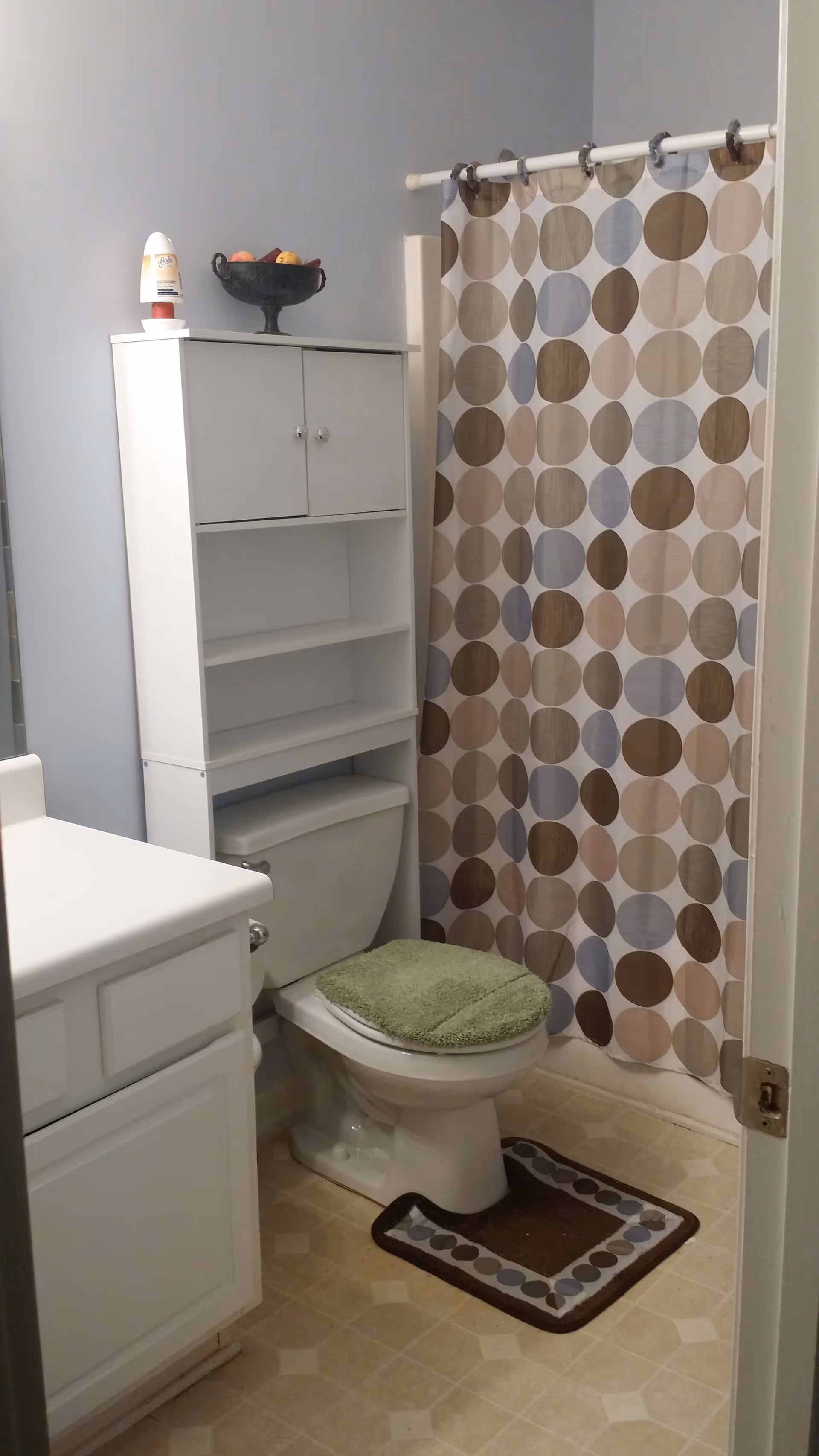 A bathroom with a white toilet that has a green cushioned seat cover and a brown and blue patterned rug in front of it. Above the toilet is a white storage cabinet with shelves and closed compartments. To the right is a shower with a curtain featuring a pattern of large circles in shades of brown, beige, and blue. To the left is a white vanity with a countertop. The floor has a beige patterned linoleum.