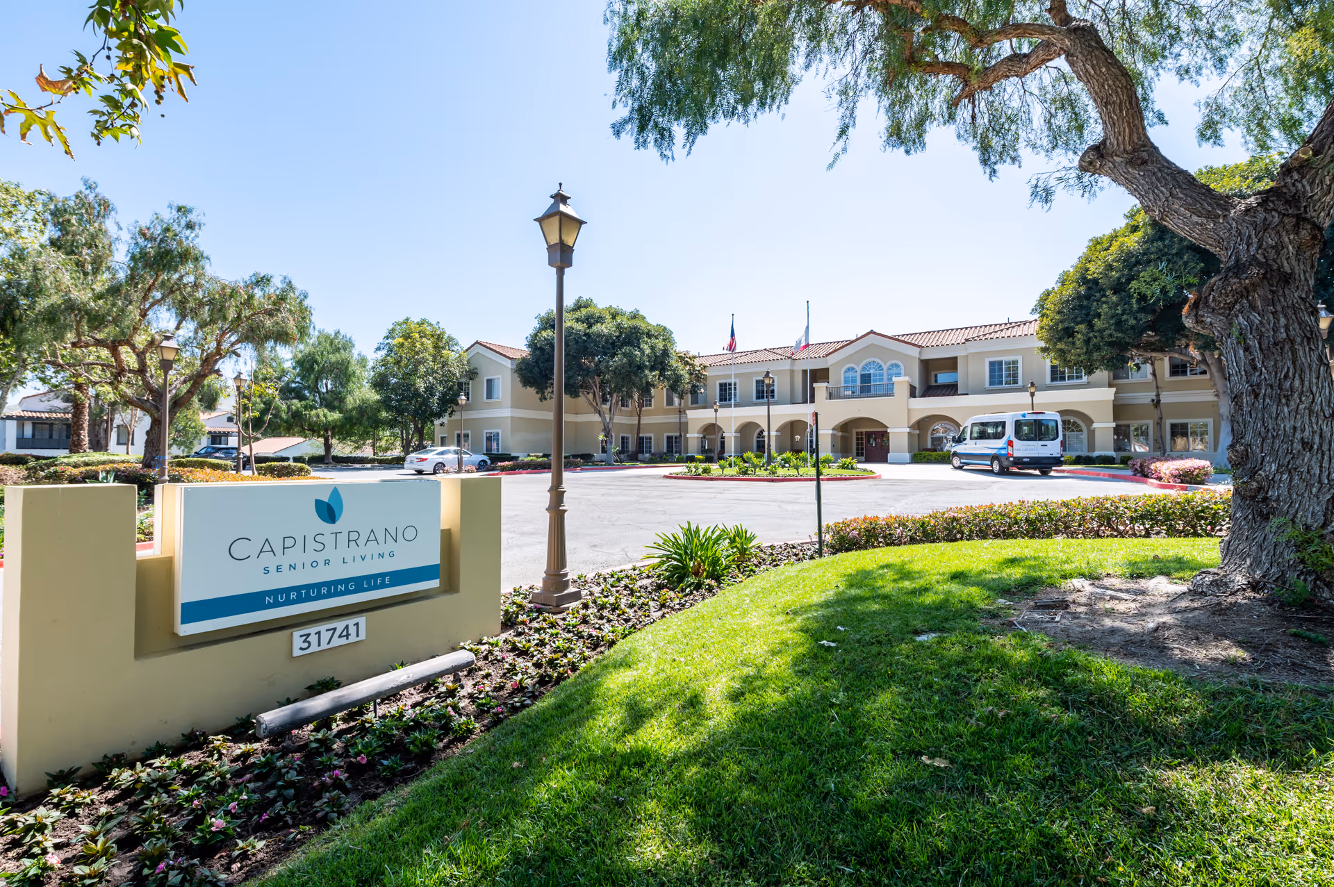 Exterior view of Capistrano Senior Living facility with a large building, well-maintained landscaping, trees, a parking area with a white van, and a sign displaying the facility name and address.