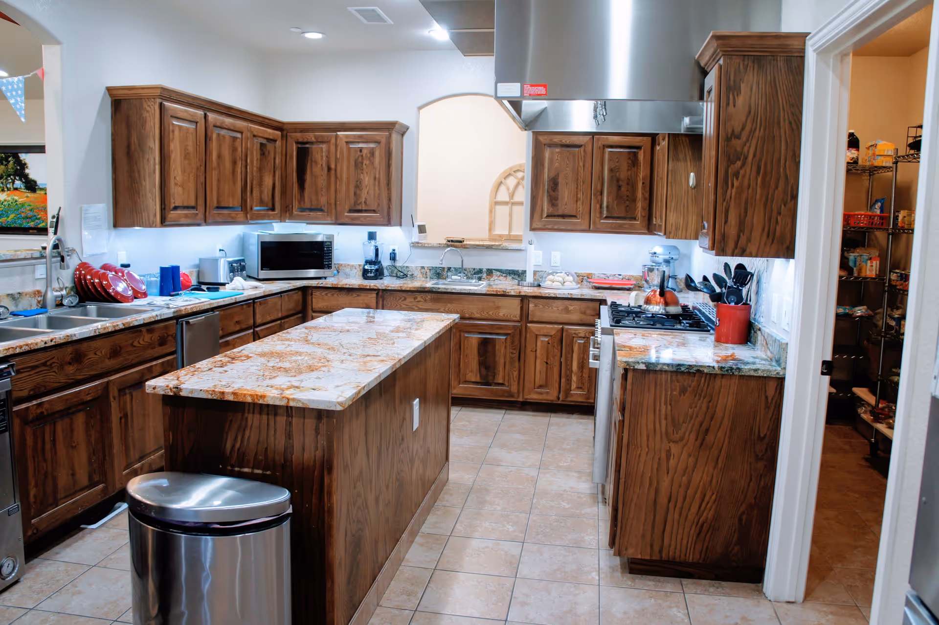 A spacious kitchen with wooden cabinets and granite countertops. The kitchen features a central island, a stainless steel trash can, a microwave, a stove with a range hood, a sink, and various kitchen utensils and appliances. There is a doorway leading to a pantry with shelves stocked with food items.