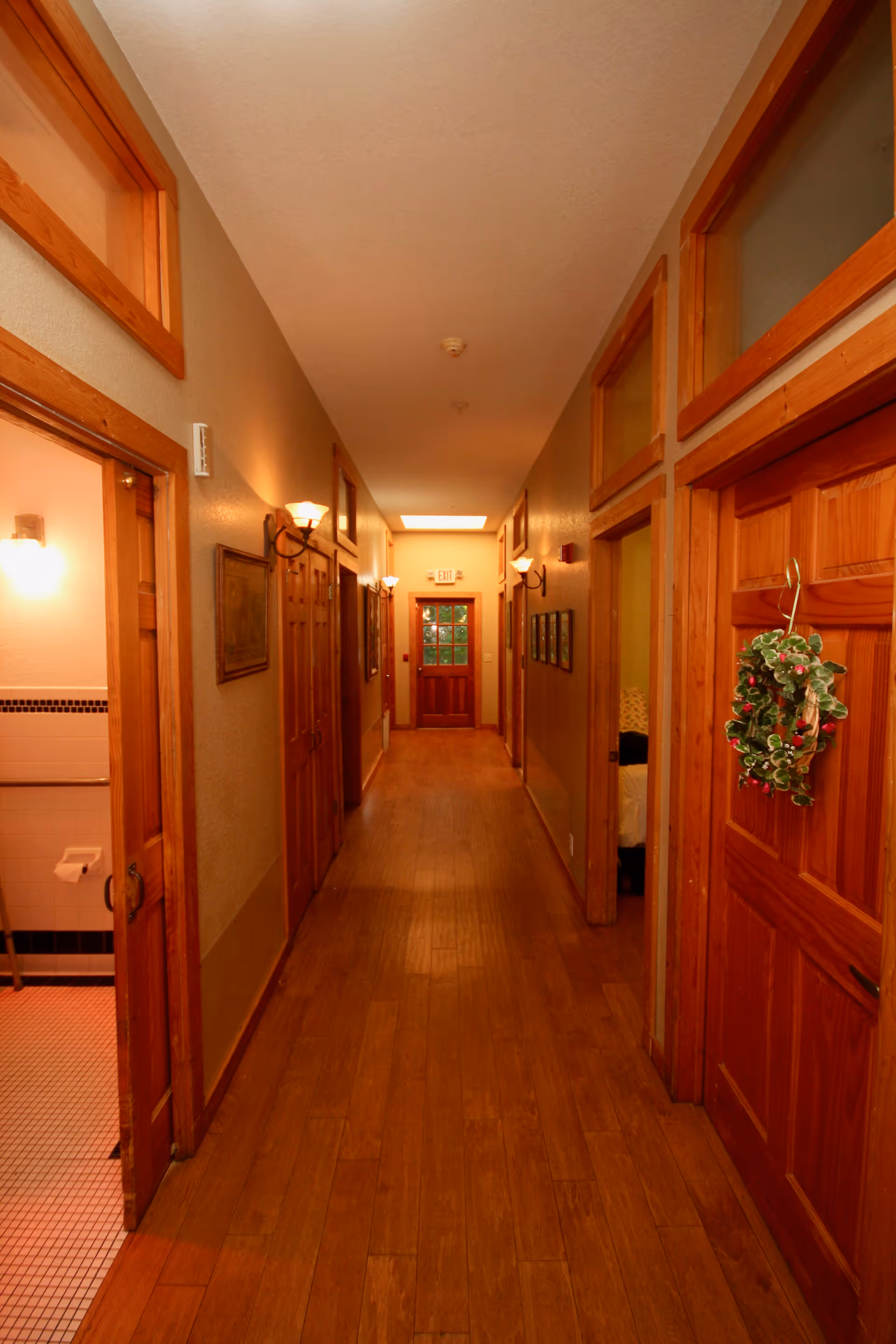 A long, narrow hallway with wooden floors and wooden doors on both sides. The walls are painted beige and have wall-mounted light fixtures. One door on the right has a decorative wreath hanging on it. The door at the end of the hallway has glass panels and an exit sign above it. A bathroom with tiled floor and walls is visible through an open door on the left.
