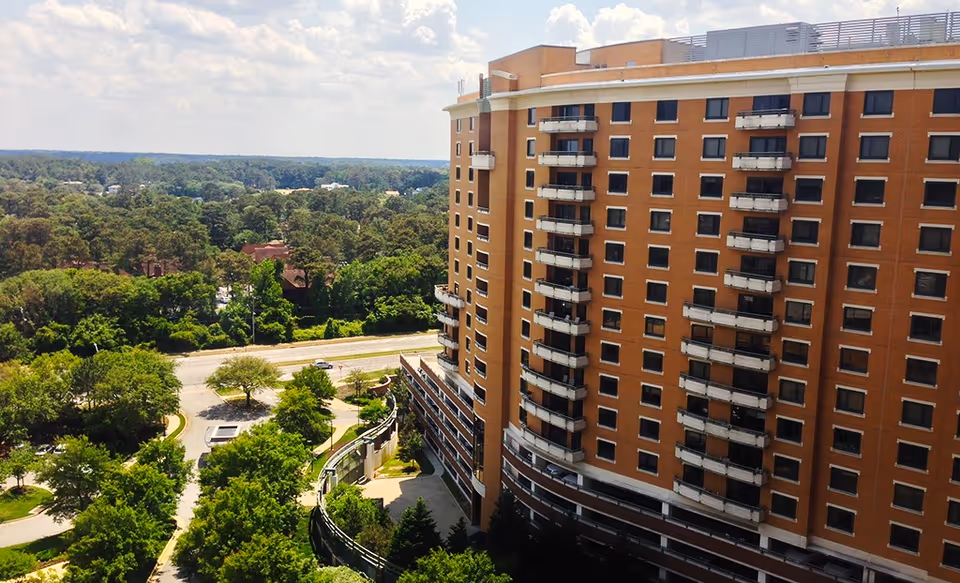 A tall multi-story brick building with balconies on each floor, surrounded by trees and greenery under a partly cloudy sky.