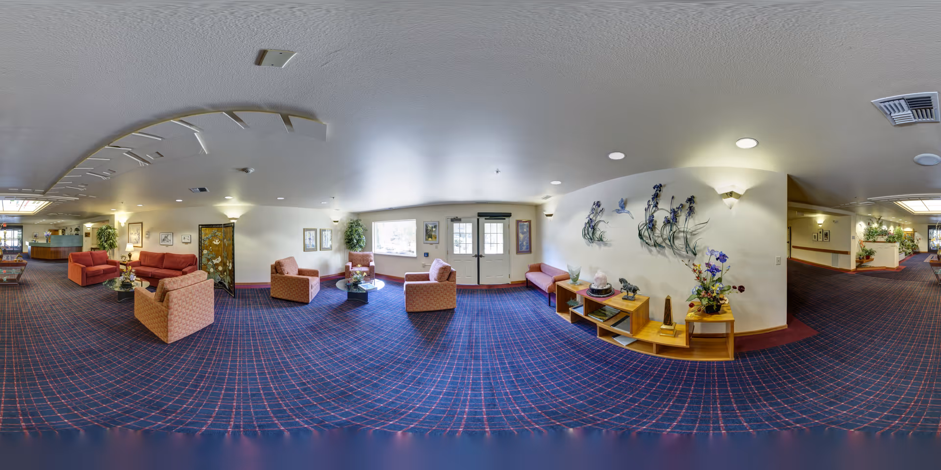 A spacious senior living facility common area with patterned blue and red carpet, multiple seating arrangements including red and beige armchairs and sofas, a glass coffee table, decorative plants, and wall art. The area is well-lit with ceiling lights and wall sconces, and there is a door with windows letting in natural light.