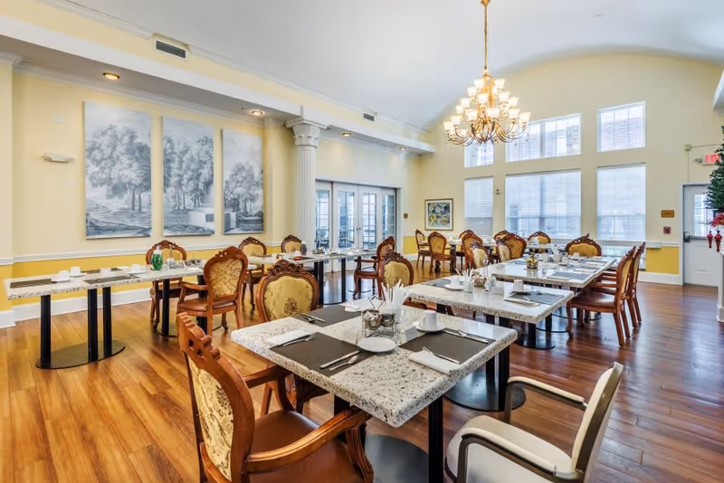 Bright dining room with multiple set tables, ornate chairs, a chandelier, and large windows.