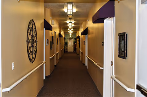 A long, well-lit hallway in a senior living facility with beige walls, handrails on both sides, decorative wall art, and multiple doorways along the corridor leading to rooms.