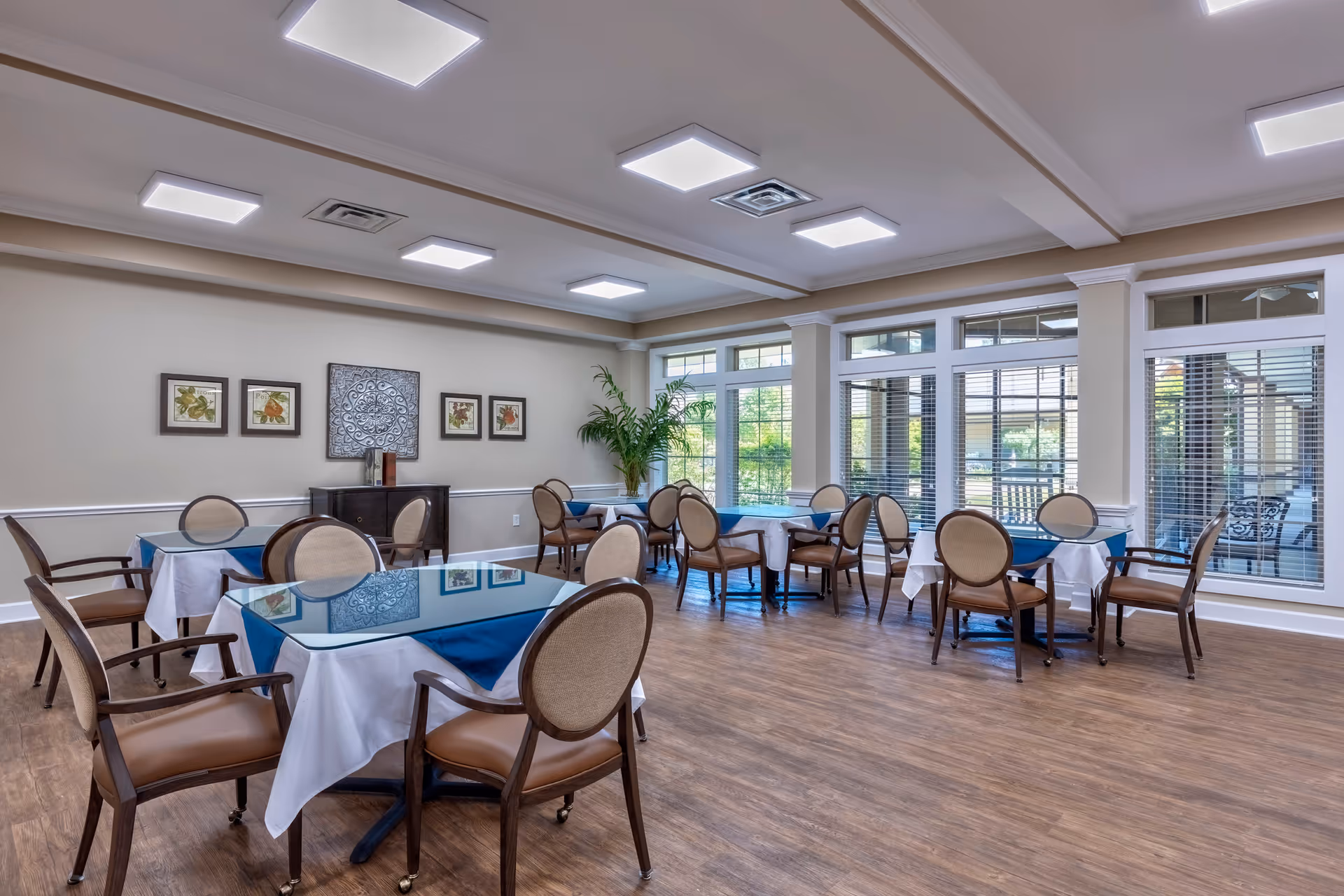 A bright dining room with several tables covered with white and blue tablecloths, surrounded by wooden chairs with beige cushions. The room has large windows with blinds allowing natural light to enter, framed artwork on the walls, and a potted plant in the corner. The floor is wooden and the ceiling has recessed lighting.