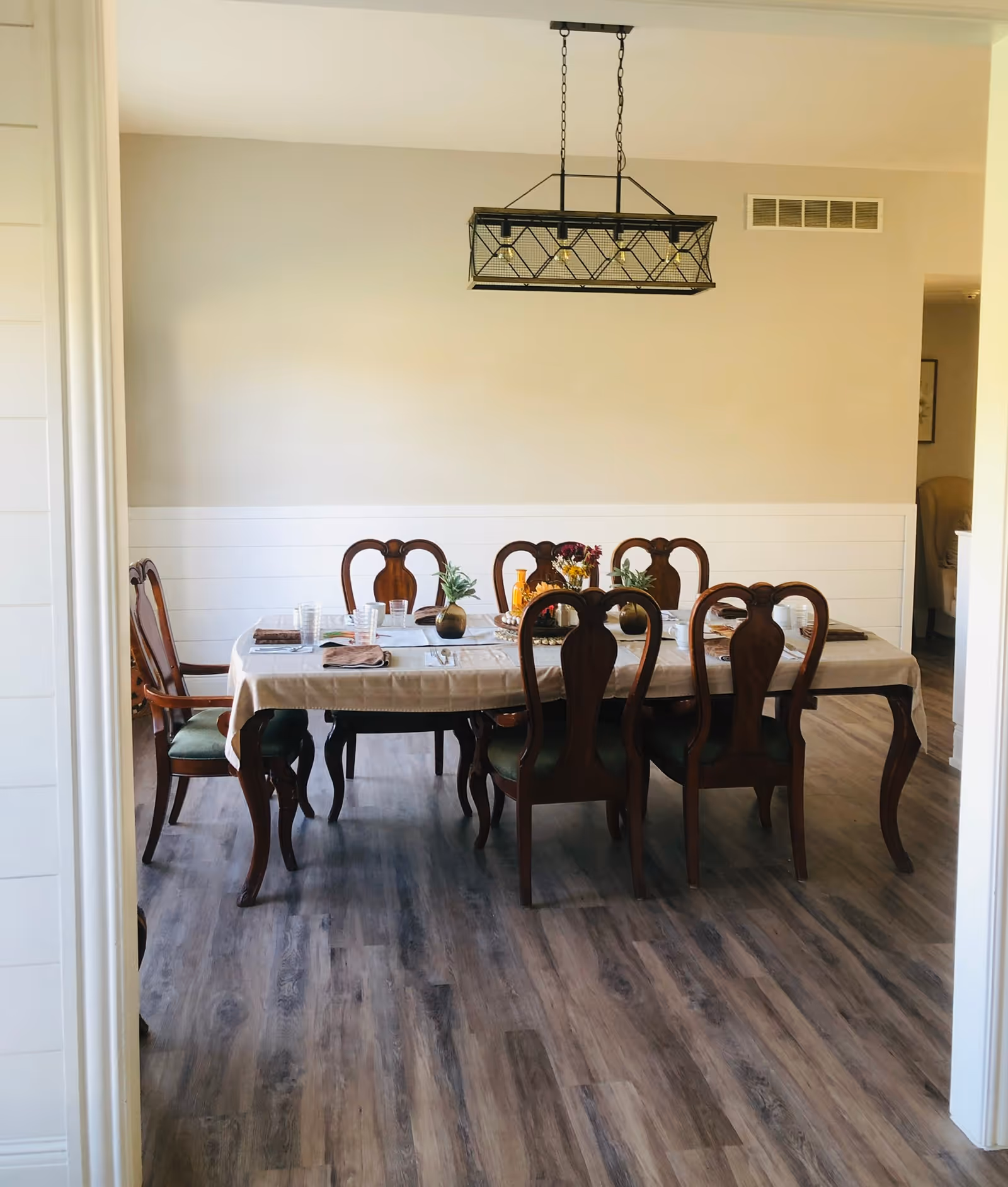 A dining room with a rectangular wooden table covered with a beige tablecloth. Six wooden chairs with green cushions surround the table. The table is set with plates, glasses, napkins, and small decorative plants. A modern rectangular chandelier with exposed bulbs hangs above the table. The room has light-colored walls and wood-patterned flooring.
