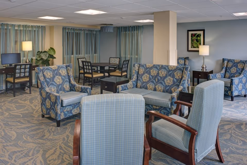 Spacious senior living common lounge with patterned armchairs, sofas, side tables, lamps, and a computer station.