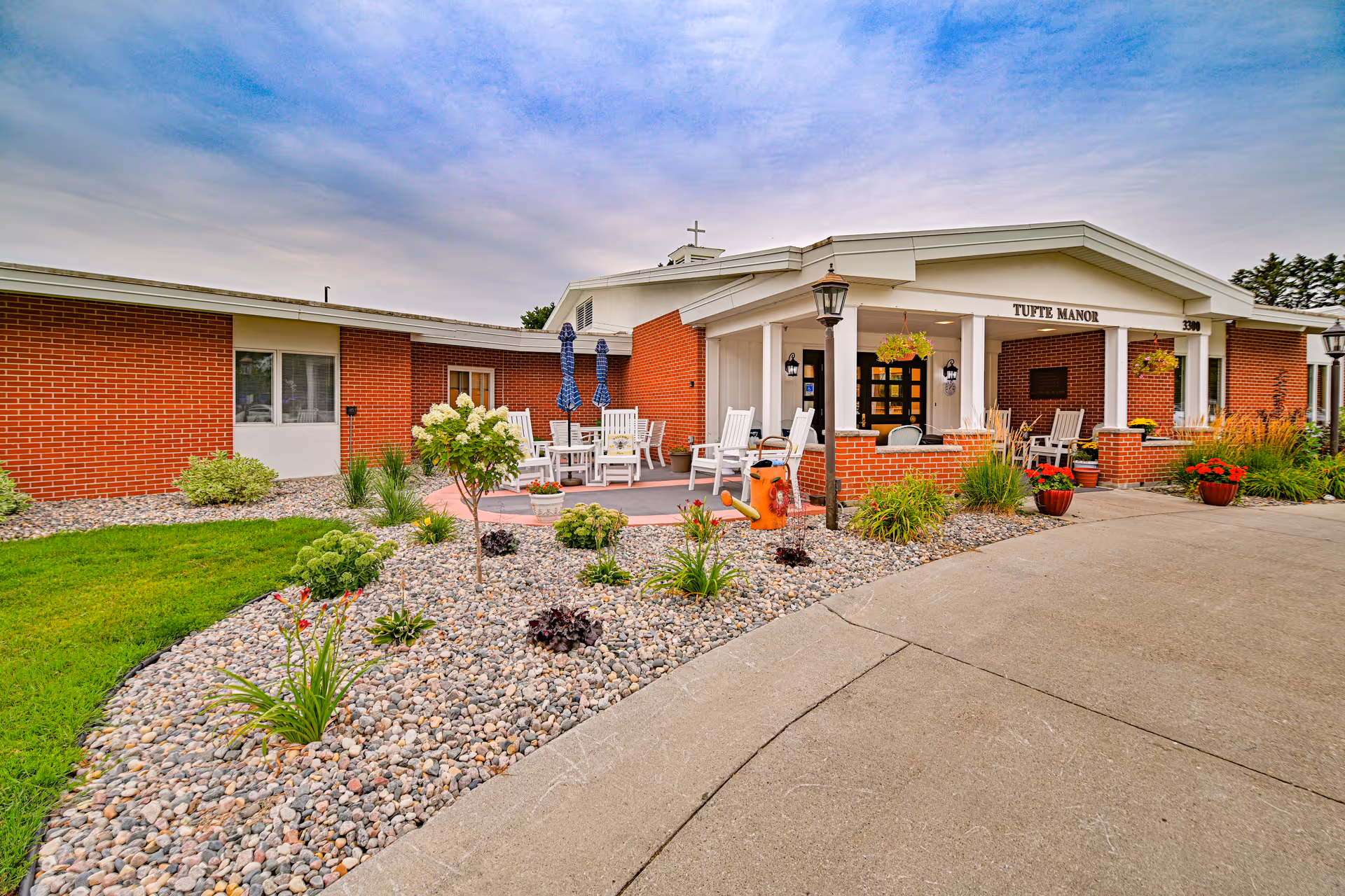 Exterior view of Valley Senior Living on Cherry - Tufte Manor, showing a brick building with a covered entrance, outdoor seating area with white rocking chairs, landscaped garden with rocks and plants, and a paved driveway under a partly cloudy sky.