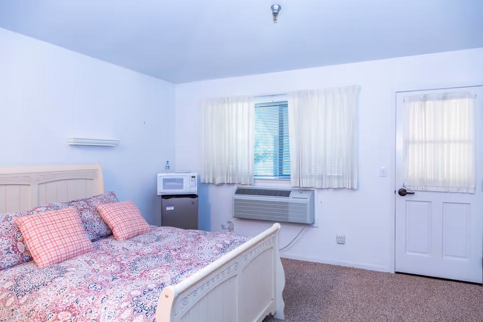 A bright bedroom with a white wooden bed frame and a bed covered with a patterned quilt and two pink plaid pillows. There is a small refrigerator with a microwave on top next to the bed. A window with white curtains and blinds is above an air conditioning unit. A white door with a window covered by a sheer curtain is on the right side of the room. The walls and ceiling are white, and the floor is carpeted.