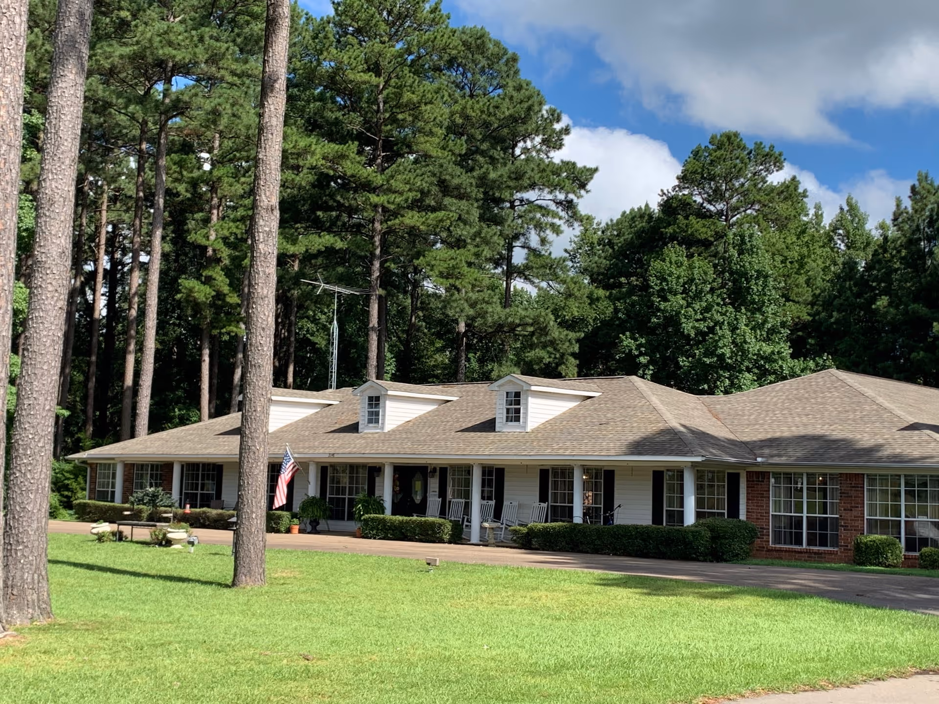 Single-story building with a beige roof and white siding, surrounded by tall pine trees and green grass under a partly cloudy sky. The building has multiple windows with black shutters, a front porch with rocking chairs, and an American flag displayed near the entrance.