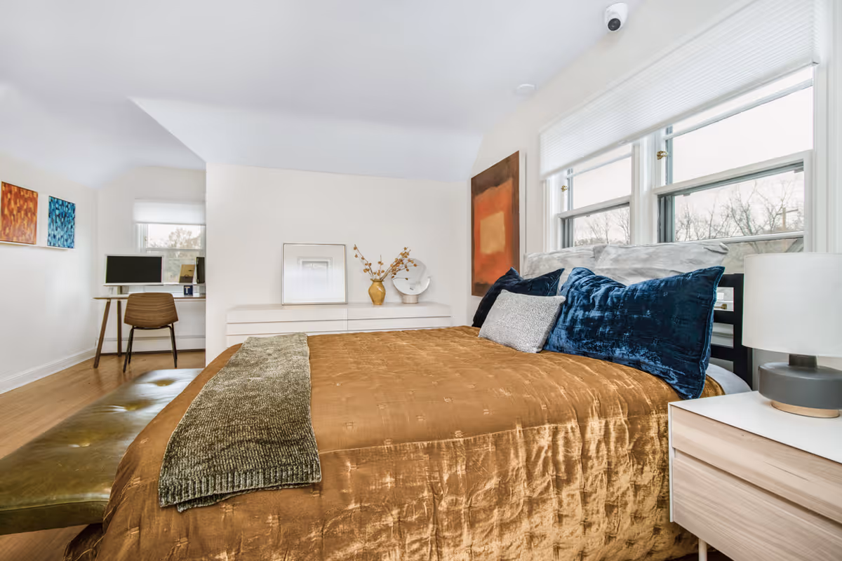 A bright bedroom with a large bed covered in a brown quilt and blue and gray pillows. There is a green throw blanket at the foot of the bed. Next to the bed is a light wood nightstand with a modern lamp. The room has three large windows with white blinds, letting in natural light. On the wall above a white dresser are decorative items including a vase with dried flowers and framed artwork. In the background, there is a small workspace with a desk, chair, and computer monitor. The walls are white, and the floor is wooden.