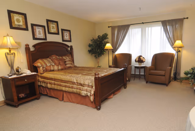 A cozy bedroom with a wooden bed frame and a neatly made bed with patterned bedding. There is a wooden nightstand with a lamp and a telephone on the left side of the bed. On the wall above the bed are four framed decorative pictures. To the right, two brown armchairs are positioned around a small round table with a floral arrangement, in front of a large window with sheer curtains. The room has beige walls and carpeted flooring, with two standing lamps and some green plants adding to the decor.