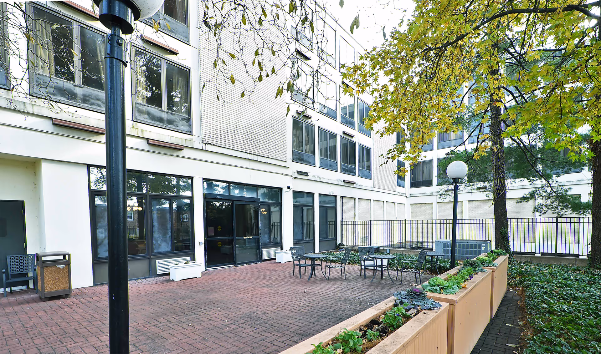 Outdoor courtyard area of Roosevelt Rehabilitation & Healthcare Center featuring brick-paved ground, several metal tables and chairs, large planters with greenery, tall trees with green and yellow leaves, and the exterior walls and windows of the building surrounding the courtyard.