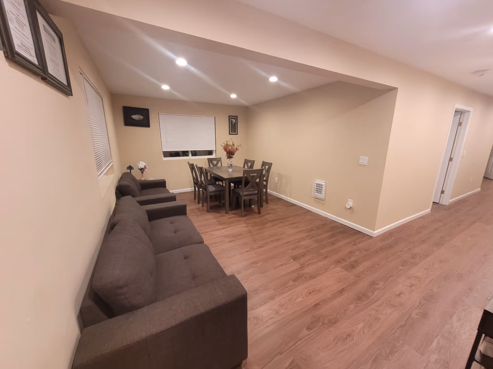 Open living and dining area with a gray sofa along the left wall, a dining table with chairs under recessed ceiling lights, and wood-look flooring.