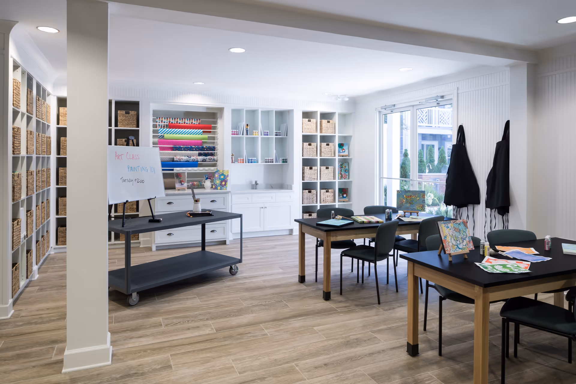 A bright and organized art room with wooden floors, white walls, and built-in shelves filled with baskets and art supplies. Two tables with chairs are set up with painting materials and colorful artwork. A rolling cart holds a whiteboard that reads 'Art Class Painting 101 Tuesday @ 2:00'. Aprons hang on the wall near large glass doors leading outside.