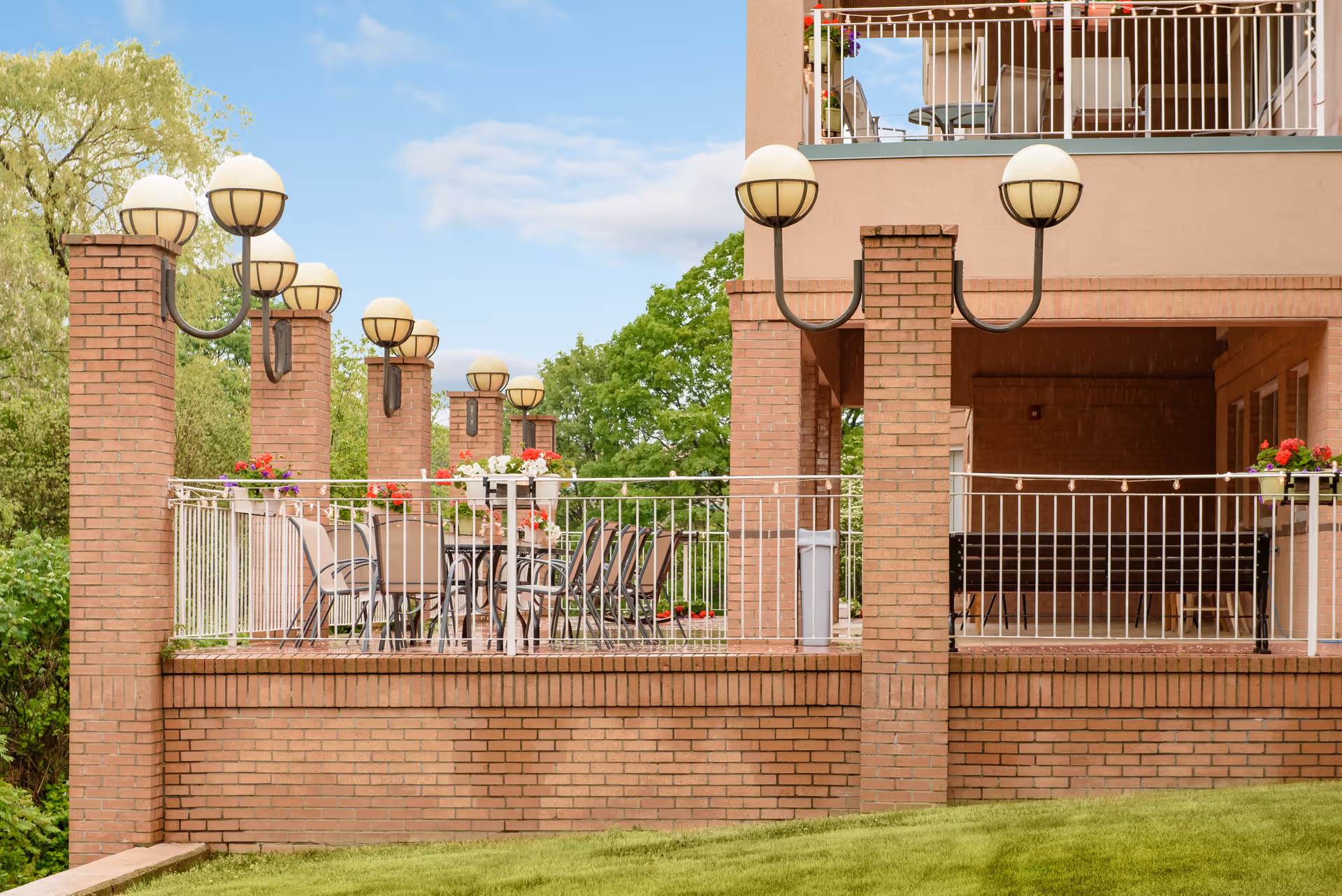 Outdoor patio area of a senior living facility with brick pillars and railings, round tables with chairs, flower pots with colorful flowers, and vintage-style globe lamps. The patio overlooks a grassy area with trees in the background under a partly cloudy sky.