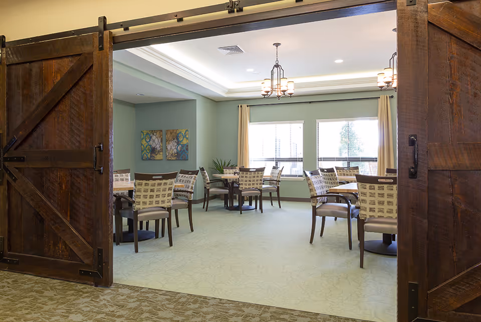 View through large wooden barn-style sliding doors into a bright dining room with several tables and chairs. The room has light green walls, two large windows with beige curtains, ceiling lights, and decorative wall art.