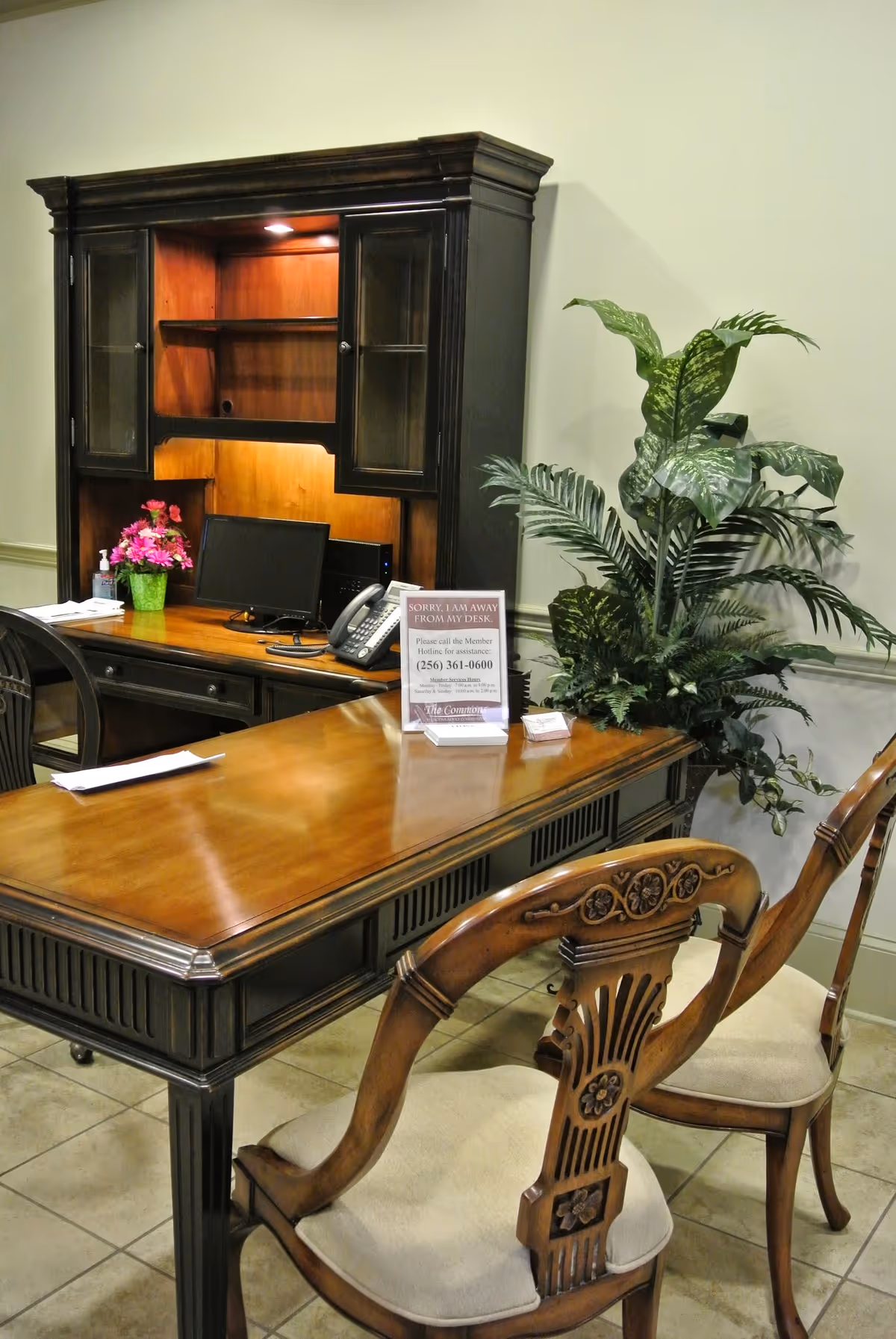 An office desk area with a wooden desk and two ornate wooden chairs with cushioned seats. Behind the desk is a dark wooden hutch with glass-paneled cabinets and shelves, a computer monitor, a telephone, and a small potted plant with pink flowers. A sign on the desk reads 'SORRY, I AM AWAY FROM MY DESK' with contact information. To the right of the desk is a large green leafy plant against a beige wall.