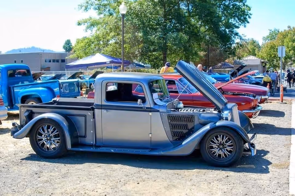 A row of vintage cars parked outdoors at a car show on a sunny day, with one classic silver and black pickup truck prominently in the foreground with its hood open. People are visible in the background walking and observing the cars.