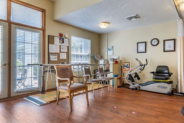 A rehabilitation or therapy room with wooden flooring, featuring a stationary exercise bike, parallel bars for walking practice, several chairs, a bulletin board with notices, and a window with blinds letting in natural light.