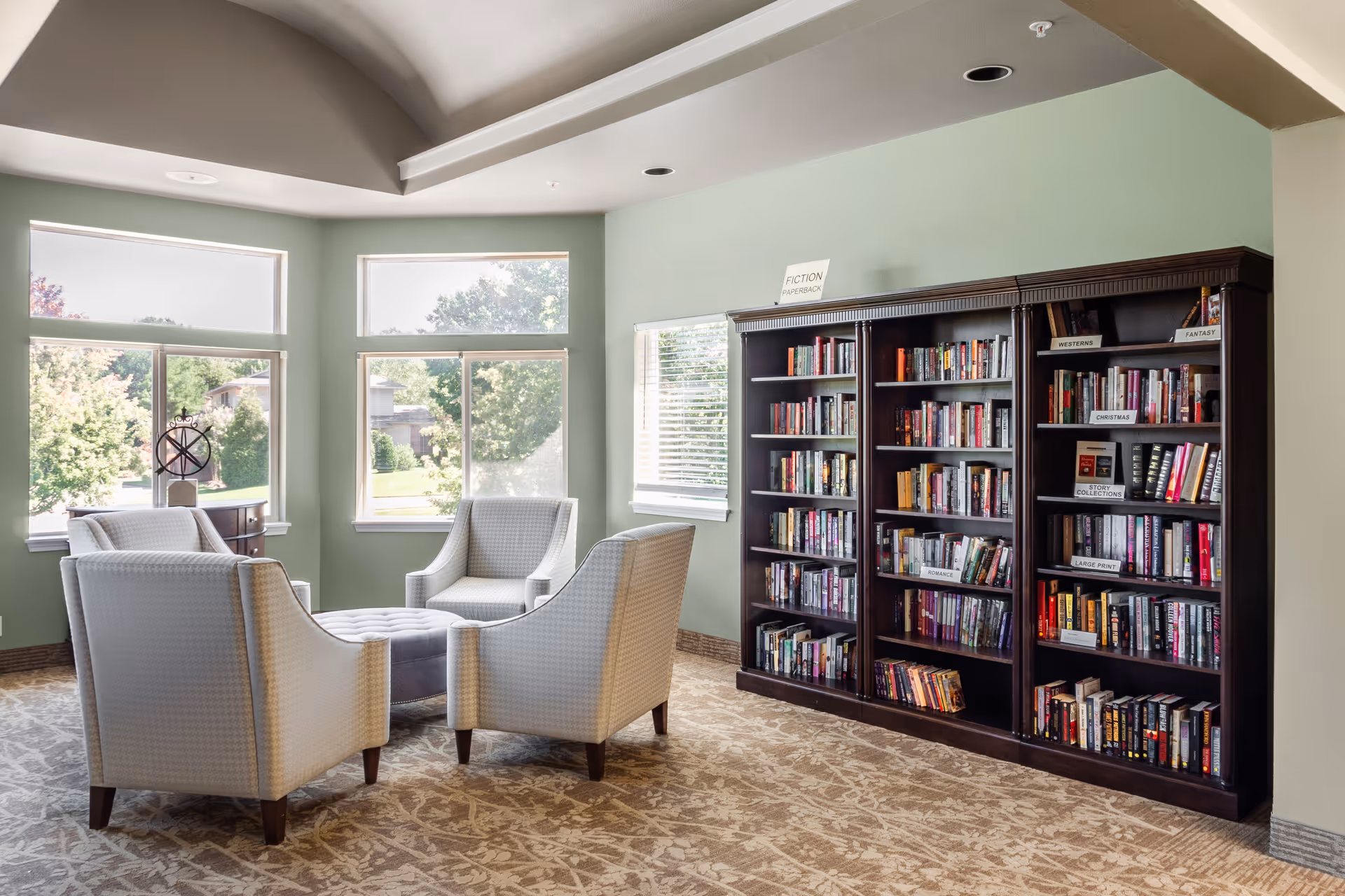 A bright communal sitting area with four upholstered chairs around an ottoman near large windows and a wall of bookshelves.