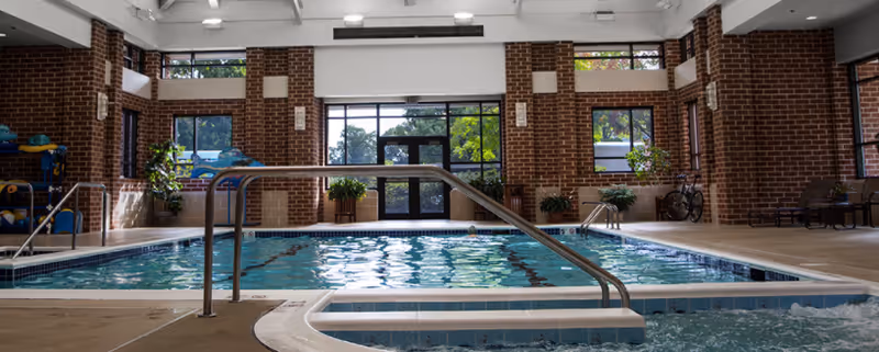 Indoor swimming pool area with clear blue water, surrounded by brick walls and large windows letting in natural light. There are handrails leading into the pool and some plants placed around the pool area.