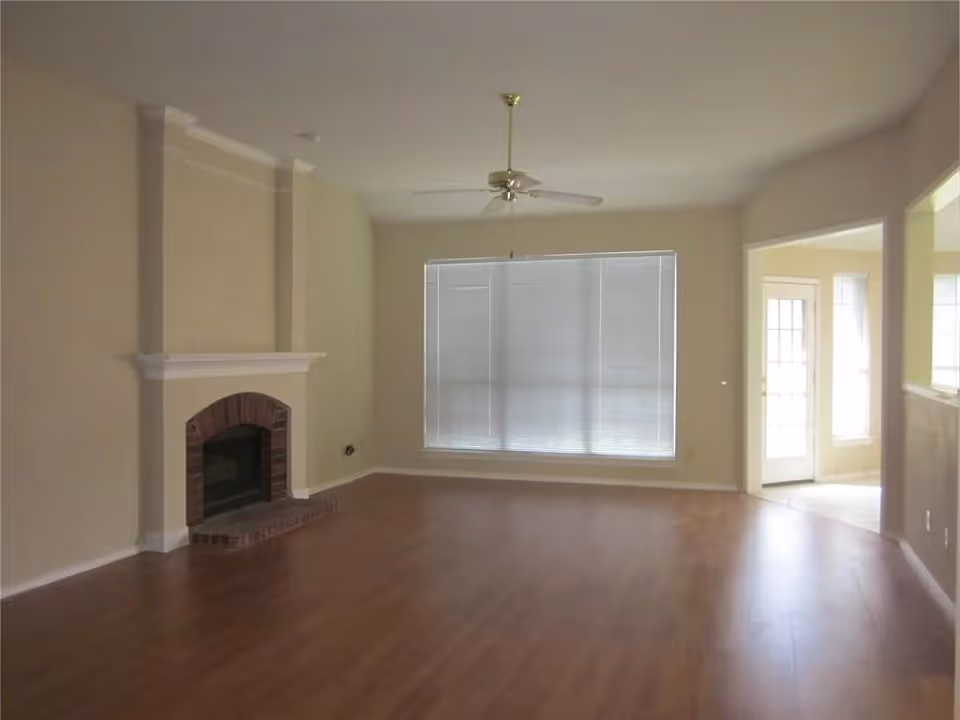 Empty living room with wooden flooring, a brick fireplace with a white mantle on the left wall, a ceiling fan, and a large window with closed blinds. There is an open doorway leading to another room with tiled flooring and glass doors.