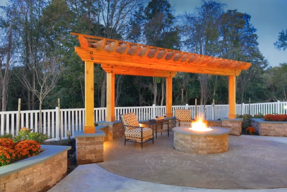 Outdoor patio featuring a wooden pergola, seating around a lit circular fire pit, and surrounding planters and fencing.