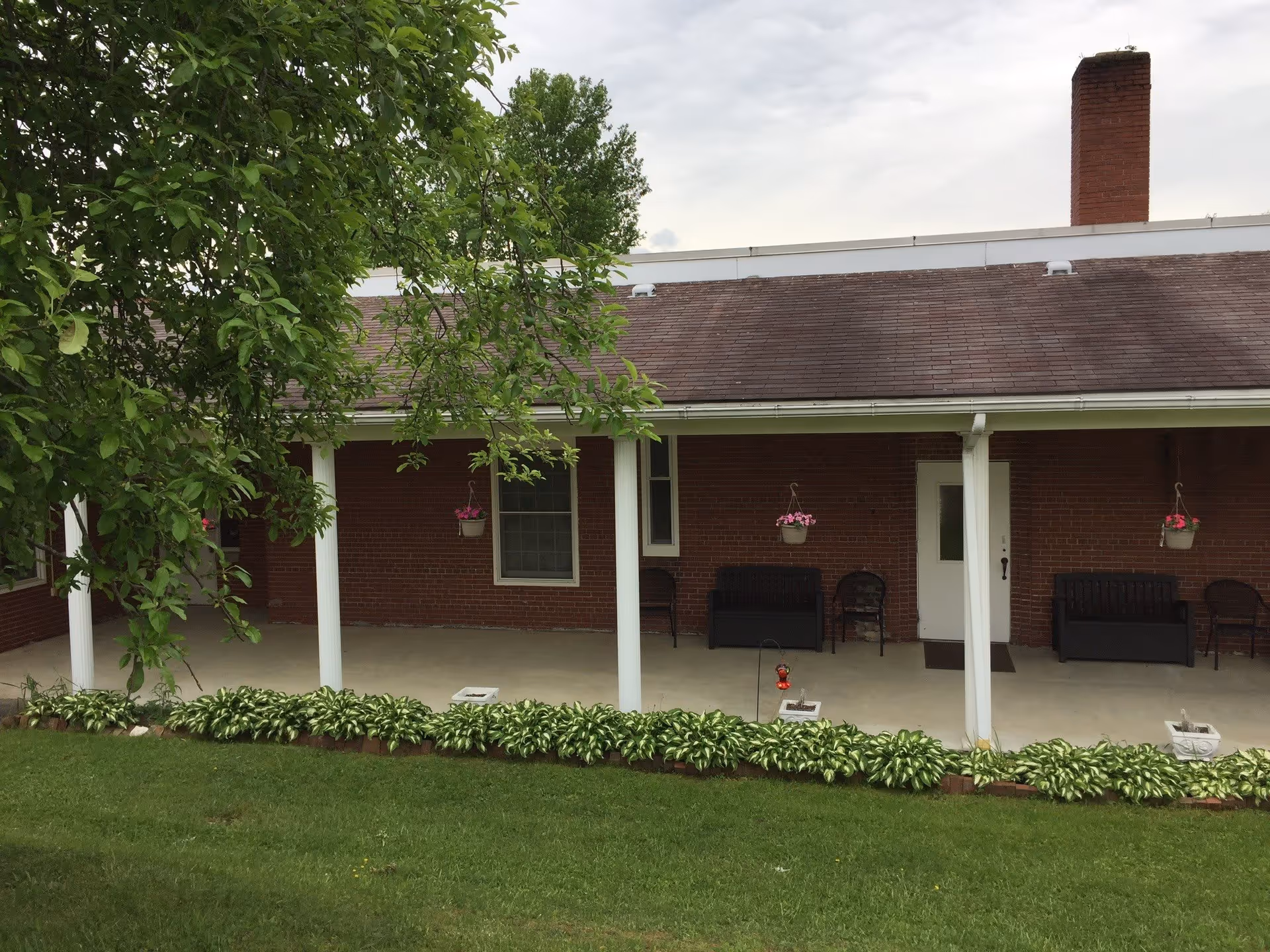 Outdoor covered patio area with white pillars, hanging flower pots, benches, and chairs in front of a brick building with a chimney. Green grass and plants border the patio.