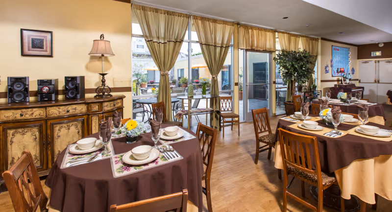 A dining room in Sharon Care Center with round tables covered in brown and beige tablecloths, set with plates, bowls, glasses, and silverware. The room has wooden chairs, a sideboard with a lamp and stereo system, large windows with beige curtains, and a view of an outdoor patio with tables and umbrellas.