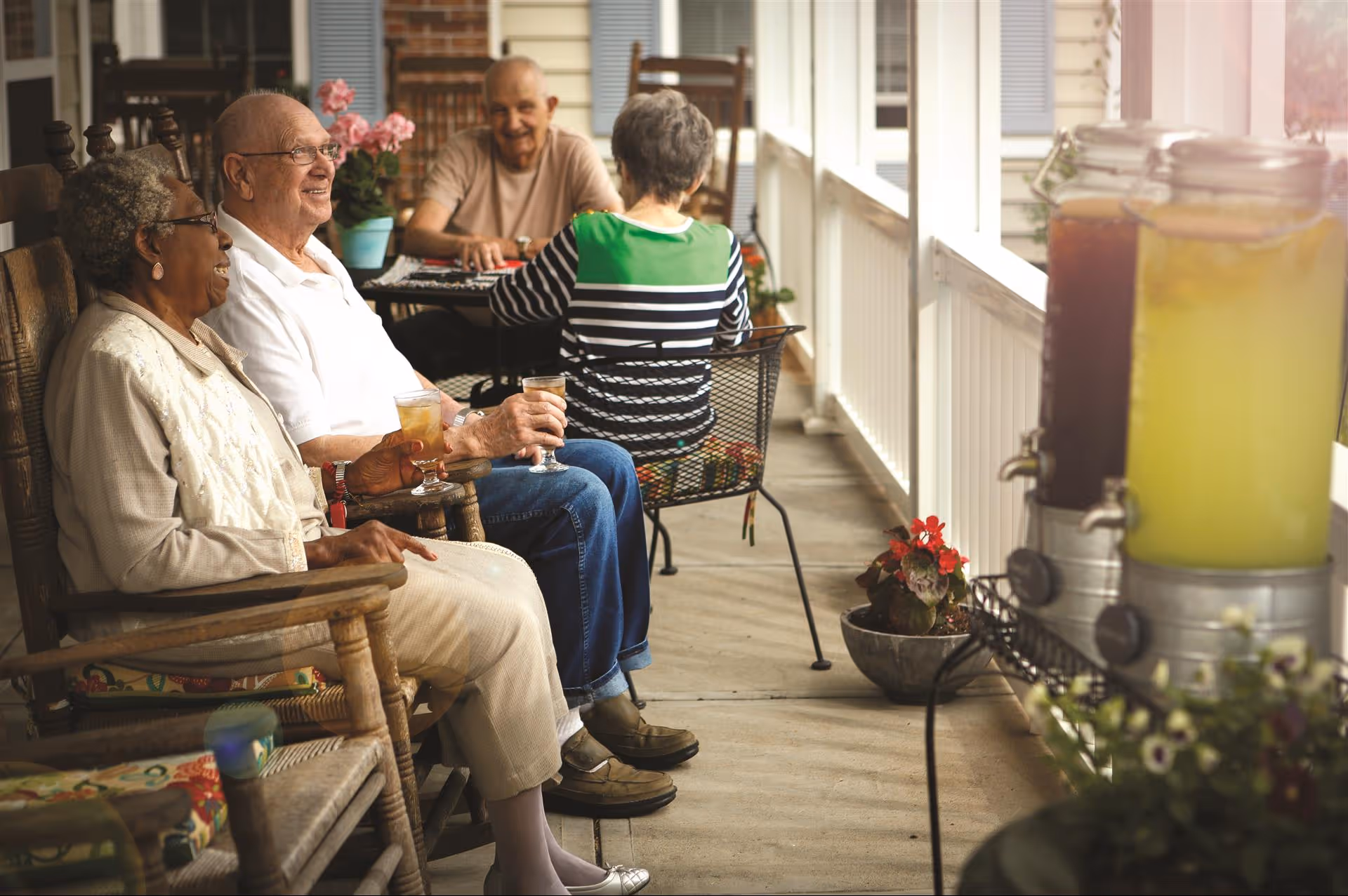 Four elderly people sitting and enjoying drinks on a covered porch. Two are seated in wooden rocking chairs in the foreground, smiling and holding glasses of iced tea. Two others are seated at a small table in the background, engaged in conversation. The porch has white railings, potted plants, and large beverage dispensers filled with iced tea and lemonade.
