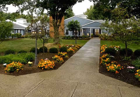 A paved walkway leads through a garden with colorful flowers and small trees towards a single-story blue building with white trim, surrounded by green grass and larger trees.