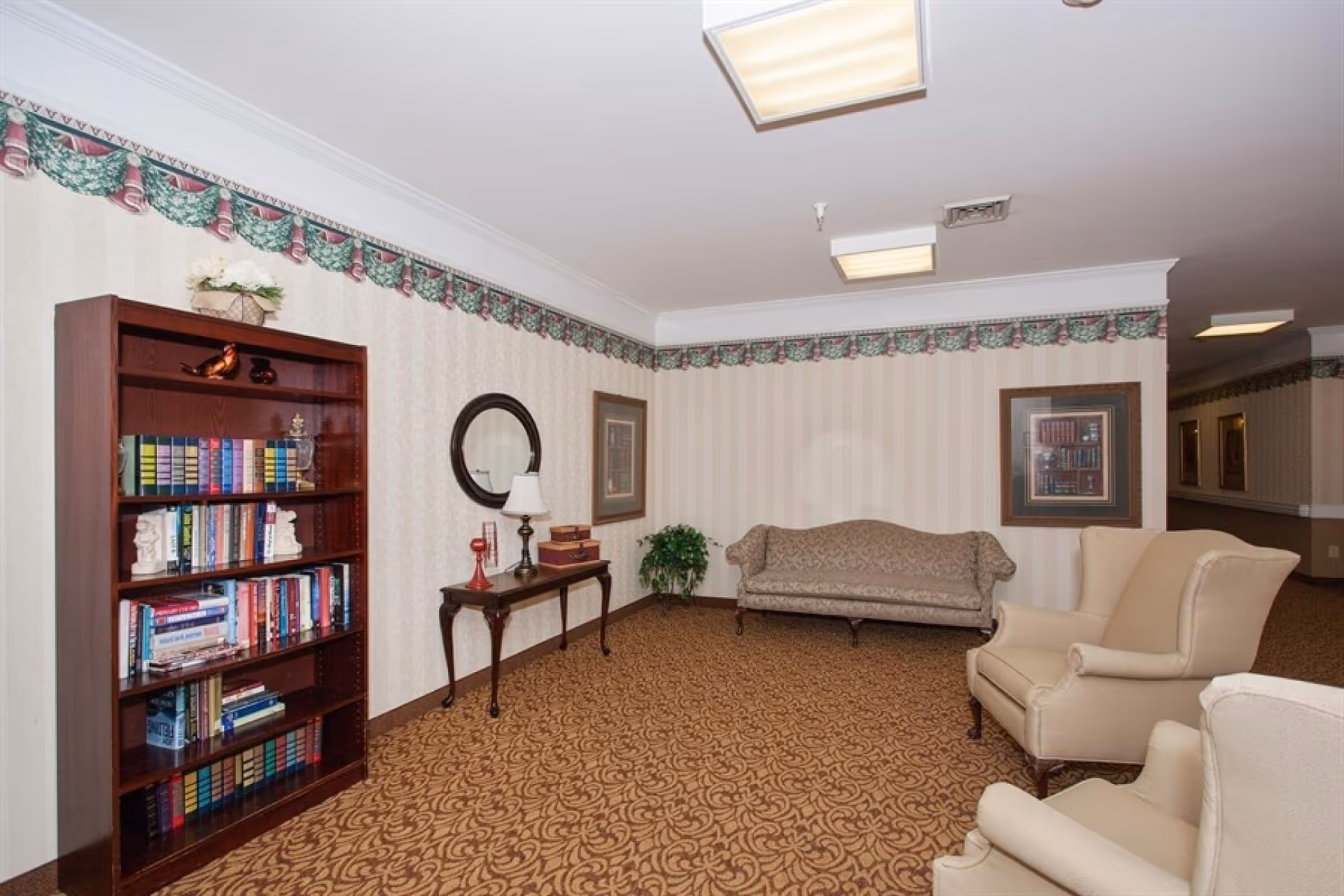Carpeted lounge area with armchairs, a sofa, a bookshelf and side table against patterned wallpaper.
