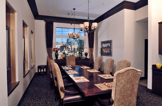 Elegant dining room with a long wooden table set for a meal, upholstered chairs, chandeliers, and a large window with drapes.