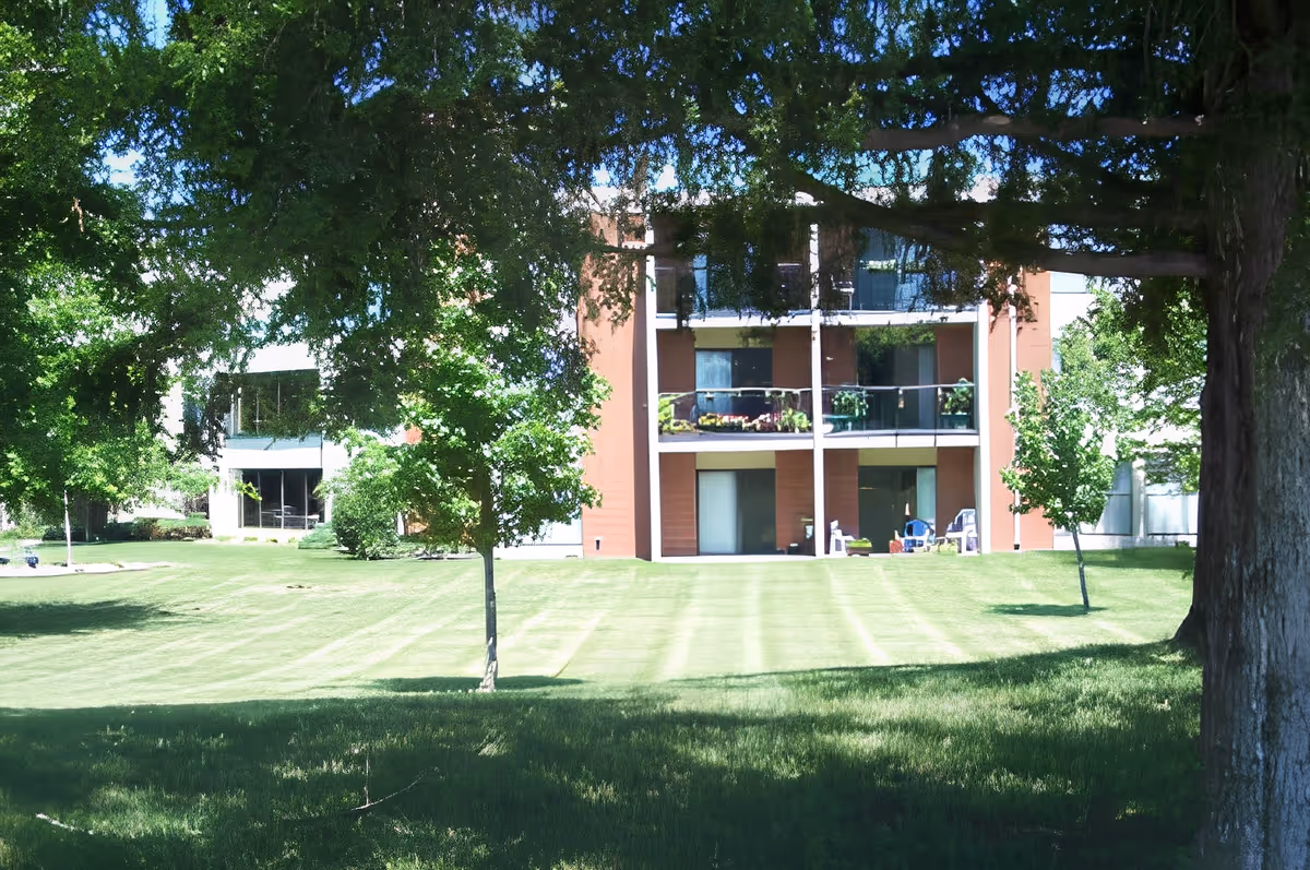 A multi-story residential building with balconies, surrounded by a well-maintained lawn and several trees providing shade on a sunny day.