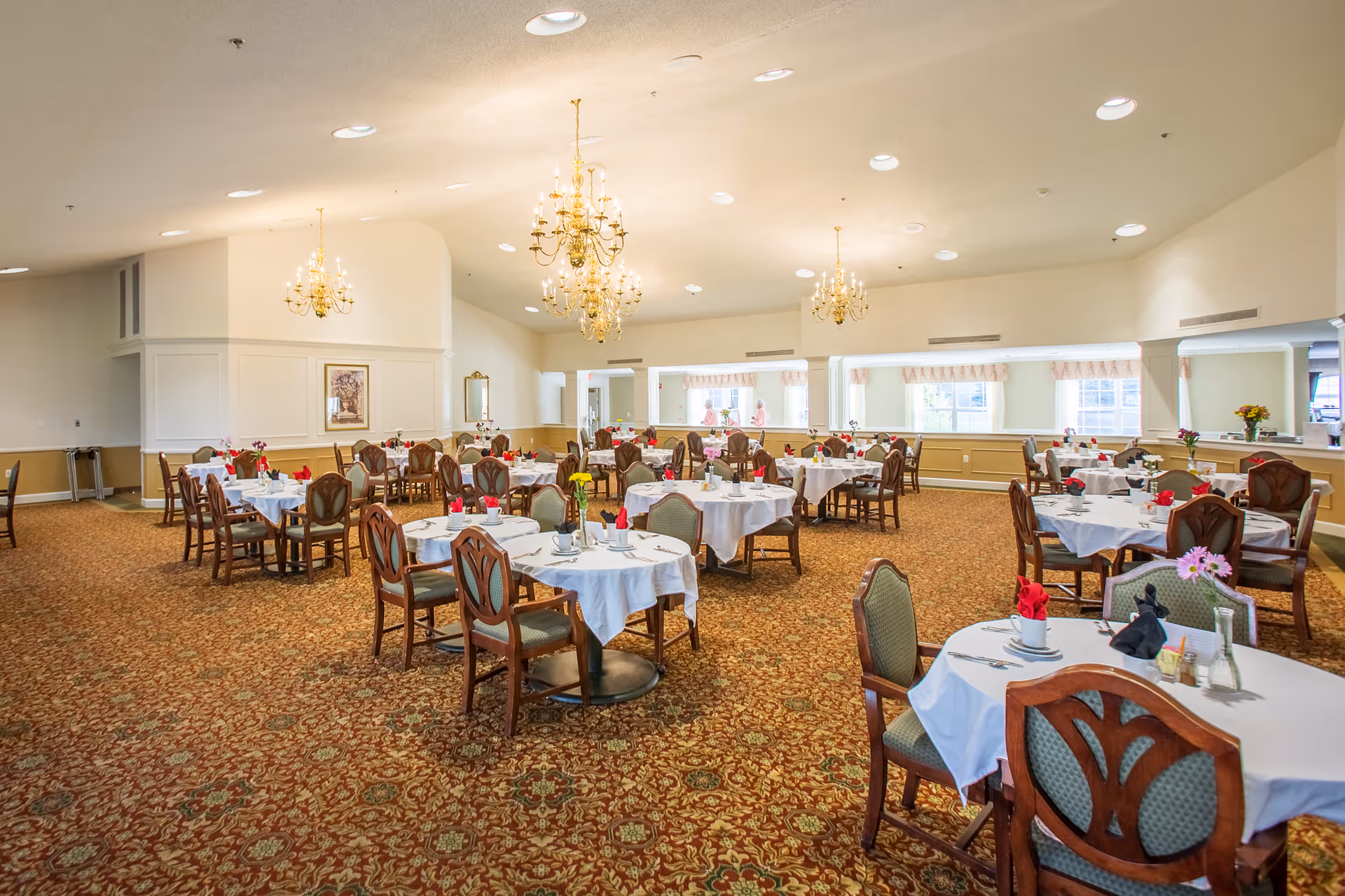 A spacious dining room with multiple round tables covered with white tablecloths, each set with cups, utensils, and napkins folded in decorative shapes. The room features ornate chandeliers hanging from the ceiling, patterned carpet, and large windows letting in natural light. There are a few people visible in the background near the windows.
