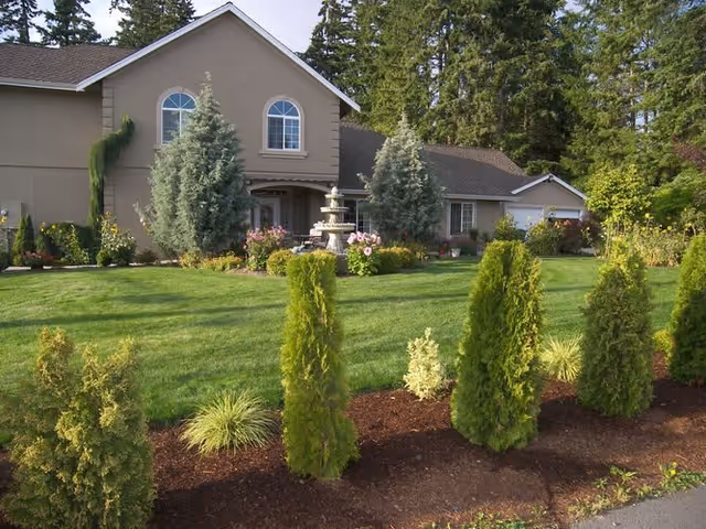 Two-story home with a manicured lawn, central fountain, and a row of young evergreen shrubs in the front yard.